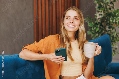 Young cheery woman wears brown shirt hold warm cup use mobile cell phone sit on blue sofa stay at home hotel flat relax spend free spare time in living room indoors grey wall. People lounge concept.