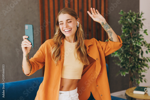 Young fun cool woman wear brown shirt listen music in earphones use mobile cell phone dancing near blue sofa stay at home flat relax spend free spare time in living room indoor. People lounge concept