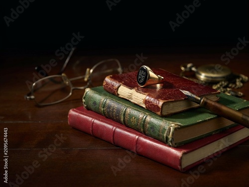 A men's signet ring with an engraved onyx stone rests on a stack of vintage leather-bound journals. Warm side lighting emphasizes the metal engraving, stone depth, and leather patina. Background featu