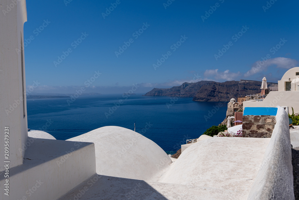 Obraz premium Traditional Cycladic architecture with smooth white domed roofs on a cliffside in Oia, Santorini. Deep blue sea and distant volcanic islands visible under a clear sky on a sunny day.