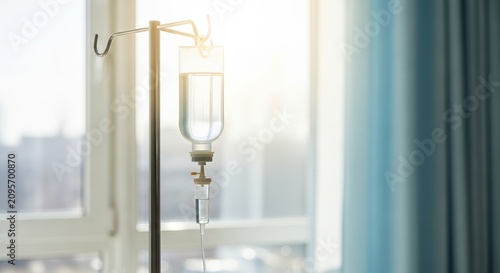 IV drip on metal stand in hospital room with natural light streaming through window. Medical equipment includes saline solution on IV stand, reflecting care and treatment atmosphere.