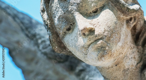 Schilderij op canvas Close-up of a weathered stone guardian angel with a sorrowful expression against a clear blue sky, symbolizing remembrance, protection, faith, eternal peace, and hope