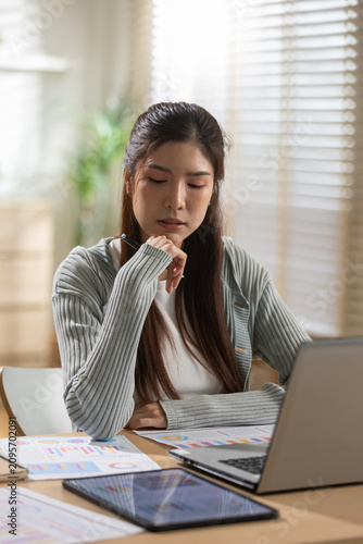 Stressed asian woman working on laptop at desk in comfortable home office reviewing document and modern tablet while thinking about difficult business finance budget problem
