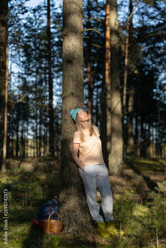 Tableau sur toile Tranquil smiling woman forager backpacker taking moment to enjoy harmony of nature in forest, smiling relaxed female leaning against tree in summer woodlands during mushroom picking hobby activity