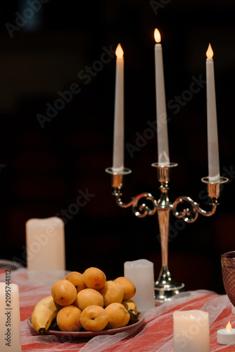 Lit white taper candles in silver candelabra with fresh yellow fruit on red tablecloth against dark background.