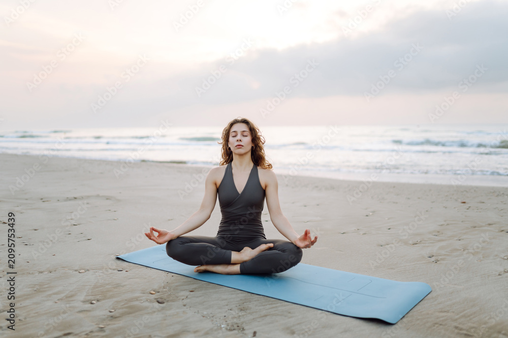 Fototapeta premium A young woman stretches on a yoga mat by the seashore. An athletic woman meditates and enjoys the seascape at sunset. Concepts of Zen, meditation, and freedom.