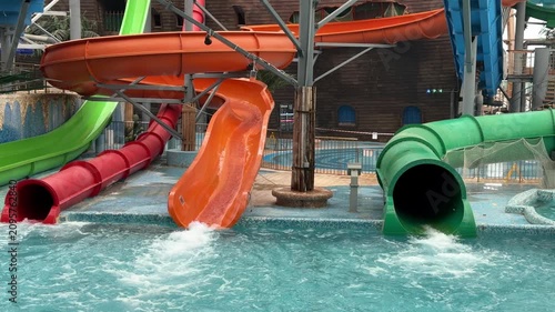  Colorful blue, orange, green, and red water slides twist into a pool inside an indoor water park with a wooden dome roof and palm trees.