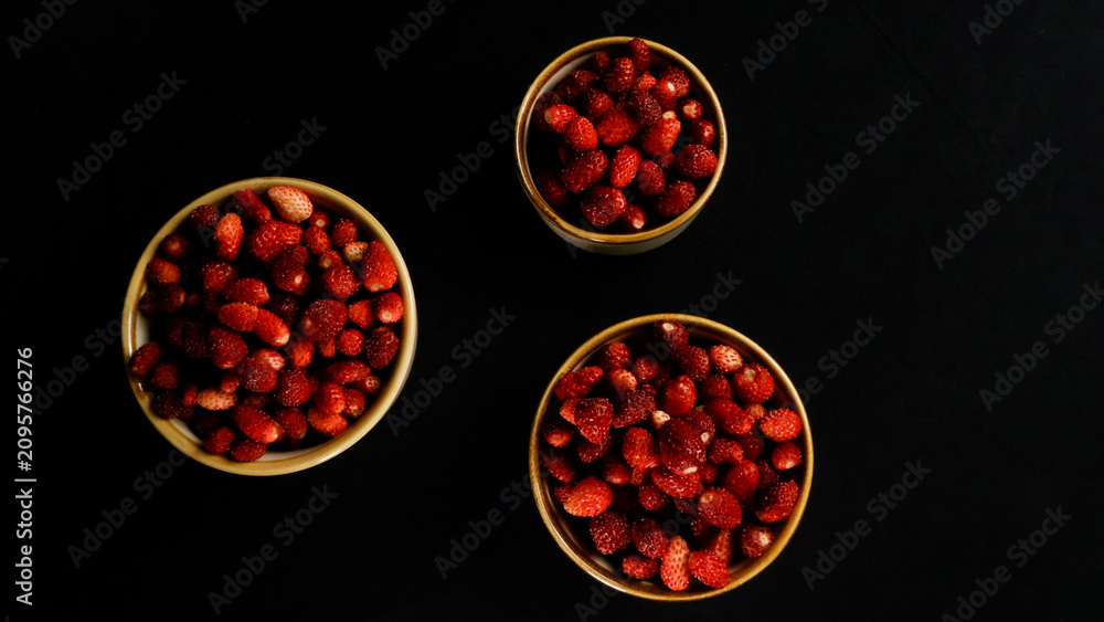Fototapeta premium Wild strawberries are arranged in three ceramic bowls. Showcasing their vibrant red color and fresh appearance against a deep black background from a top down perspective