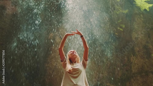 A happy blonde woman in a light t-shirt reaches out to touch a powerful waterfall in a lush tropical forest during daytime, enjoying the cascading water and natural beauty.