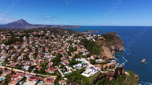 Aerial view of white villa homes along dramatic cliff coastline near Javea, Mediterranean Sea and rocky coves visible with Montgo mountain peak in distance, Alicante, Spain