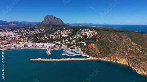 Aerial view of Javea marina with docked sailboats, turquoise Mediterranean waters, coastal town buildings, rocky cliffs and the iconic Montgo mountain peak, Costa Blanca, Spain