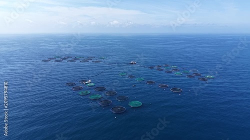 Aerial view of an offshore fish farm featuring clusters of circular aquaculture cages connected by towlines, with a service vessel operating among the pens and open ocean horizon