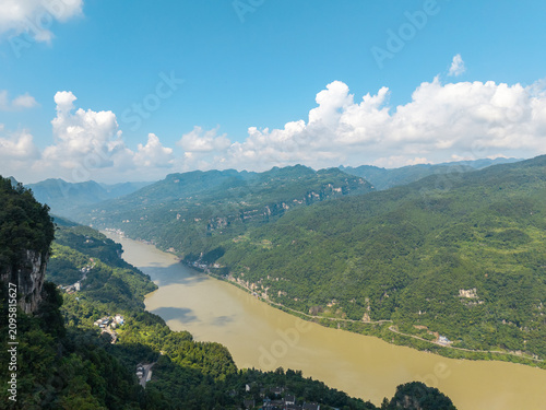 The scenic landscape shows the Three Gorges Reservoir winding through the lush green mountains near Yichang in China under a bright blue sky with white clouds