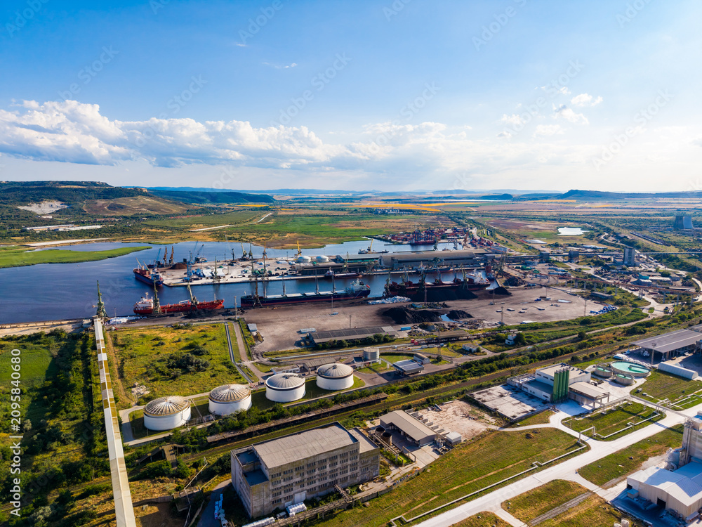 Fototapeta premium View from above shows a large industrial site with cranes, ships, and storage tanks near a river and open land during the daytime