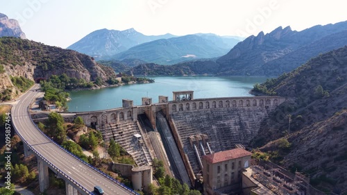 Aerial view of Oliana dam on the Segre River with elevated highway bridge curving through valley, turquoise reservoir and jagged mountain peaks, Lleida, Catalonia, Spain
