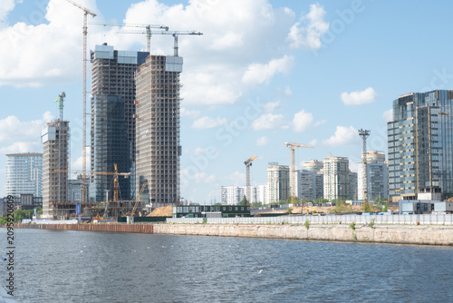 Wide horizontal view of new residential towers under construction with multiple cranes, located on a concrete river embankment. Ideal for urban planning, architecture design, and city landscape