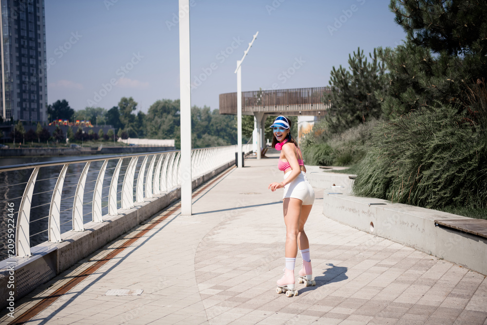 Fototapeta premium Young woman roller skating on a sunny waterfront promenade wearing retro outfit and visor, smiling and looking back while enjoying outdoor fitness and leisure