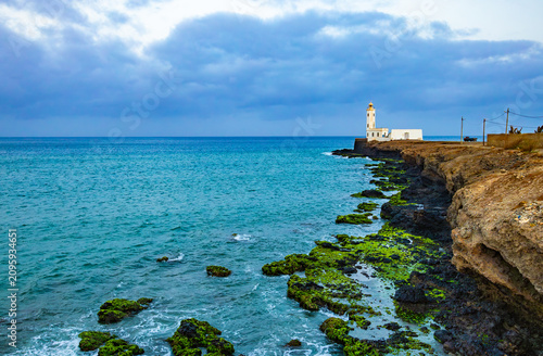View of Dona Maria Pia lighthouse on rocky volcanic coastline with green mossy stones and turquoise Atlantic ocean under cloudy sky in Cabo Verde