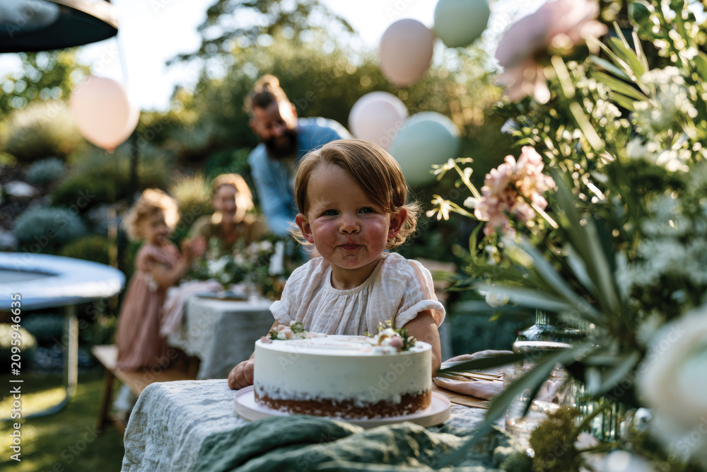Fototapeta premium A happy child with short brown hair is celebrating a birthday party outdoors, surrounded by family, enjoying a beautiful cake on a sunny day in the garden.