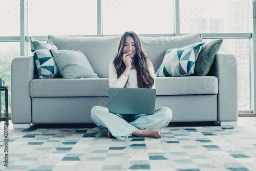 Young woman working on laptop at home sitting on floor in front of sofa  smiling and relaxed in bright living room with modern pillows and rug