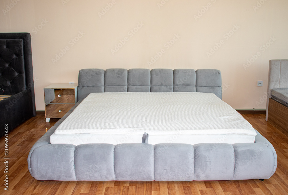 Obraz premium Modern king size double bed with white mattress and quilted grey velvet frame displayed in a furniture showroom interior front view.