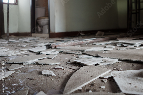 A low-angle view of scattered debris, broken ceramic tiles, and construction waste covering the floor of an indoor building space during renovation, with an open doorway leading to a restroom in the b