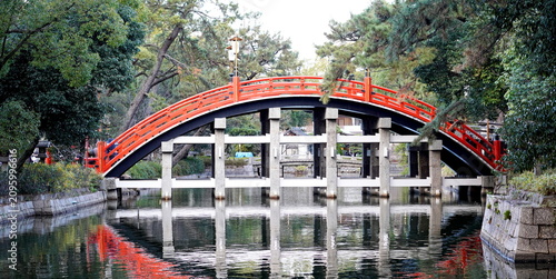 Slika na platnu 日本の伝統的な庭園に架かる鮮やかな朱色の太鼓橋が、穏やかな水面に美しく反射している風景。周囲の豊かな緑の木々が、静かで落ち着いた和の空間を創り出しています。