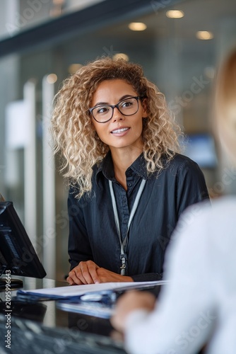 A female bank worker talks to a client at the counter. The setting is a bank during working hours. Both are focused on the conversation about banking services