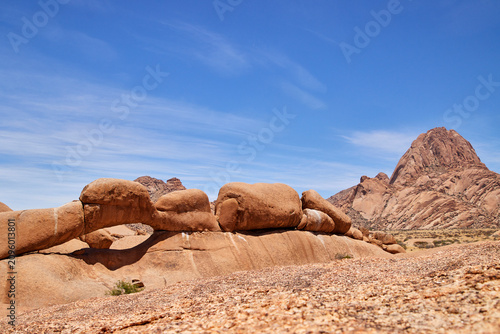 Konstfotografi Ein natürlicher Granitbogen liegt vor einer markanten Berglandschaft unter blauem Himmel an der Spitzkoppe in Namibia
