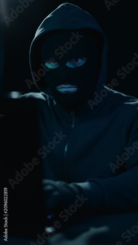 Hacker in hood and balaclava at laptop in dark room