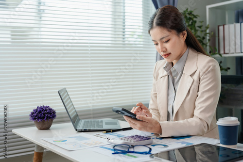 Professional Asian businesswoman talking on smartphone while looking at laptop and holding pen at her office desk with documents.