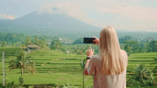 A blonde woman photographed from behind taking a picture of a volcano and rice terraces on a bright sunny day. She stands facing the scenic landscape outdoors.