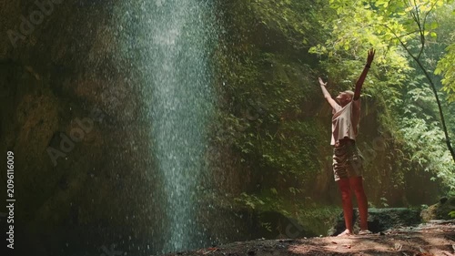 A happy young woman joyfully raises her hands to the sky near a fast-flowing waterfall surrounded by lush tropical vegetation on the island of Bali.