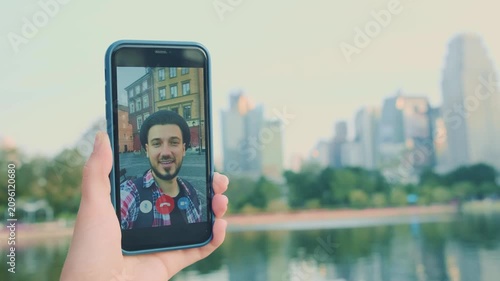 A female hand holds a phone showing a man speaking on a video call. In the background is Bangkok's business center with skyscrapers and a lake in a park.