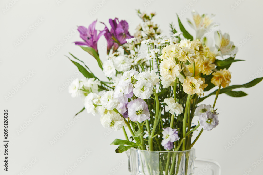 Obraz premium Fresh mixed bouquet of purple alstroemeria and white yellow stock flowers in a clear glass jug. Spring floral arrangement on a light background.