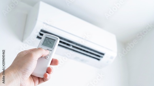 A hand holds a white plastic remote control to adjust the temperature settings on a modern white wall air conditioner