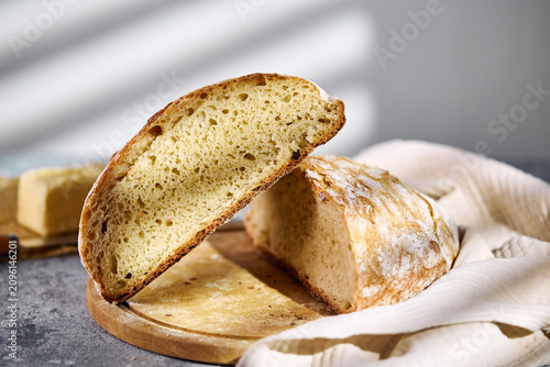 Close-up of a freshly baked artisan loaf cut in half, revealing porous crumb structure. Stacked on a wooden board with butter and linen cloth nearby.