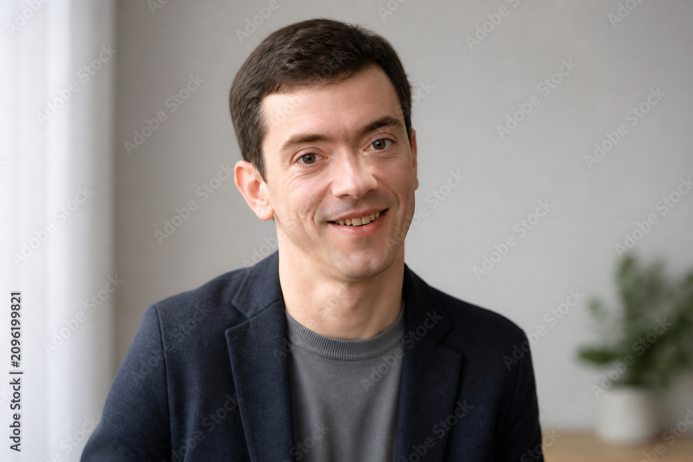 Fototapeta premium A smiling man in a dark blazer is posing indoors against a light background.