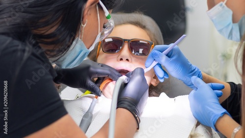 фотографія A young girl with light brown hair and sunglasses smiles in the dentist's chair as dental professionals tend to her