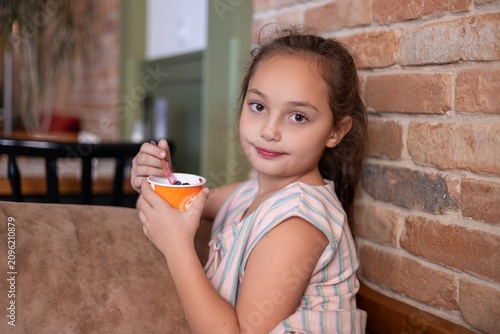 A young girl with brown hair enjoys a bowl of ice cream, smiling softly while seated in a cozy café setting.