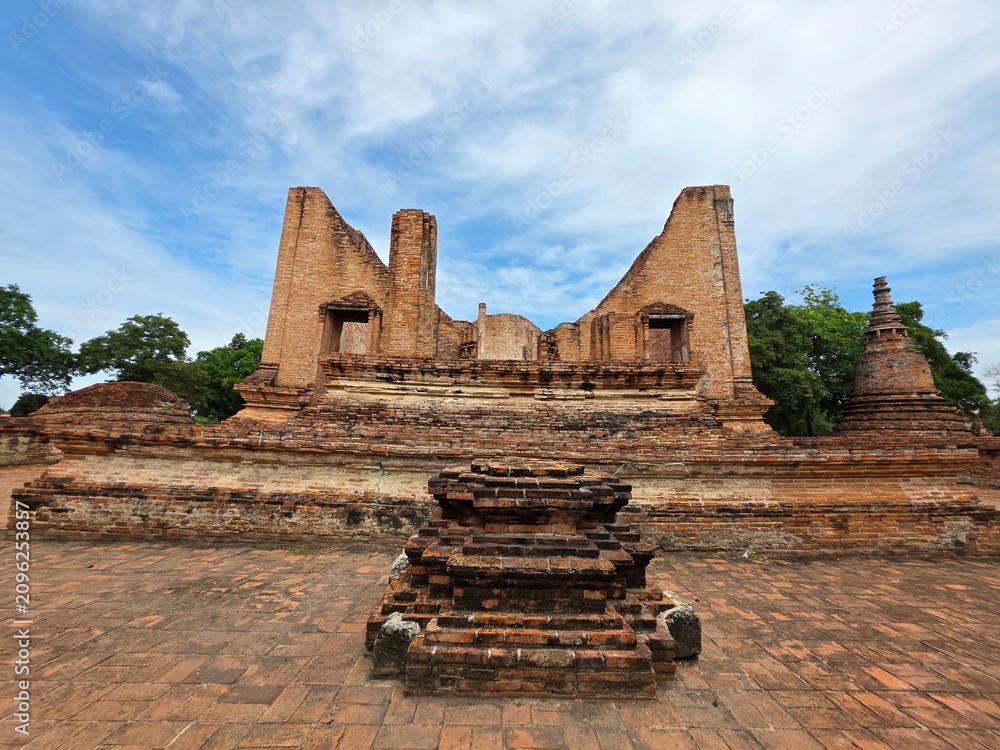 Fototapeta premium Ancient Ruins in Thailand: Sun-drenched ancient ruins in Thailand, featuring weathered brick structures and pagodas, stand testament to a bygone era under a clear blue sky dotted with fluffy clouds.