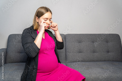 Pregnant woman sitting on a sofa while wiping away tears, expressing emotional stress, sadness, or pregnancy mood swings. Concept of prenatal mental health, emotional well-being, hormones 