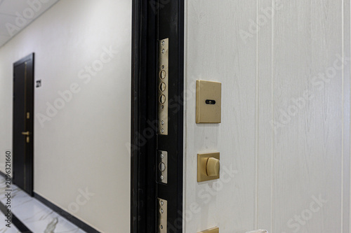 black metal entrance door features brushed‑steel lock cylinders and hardware. beside it, beige wall holds doorbell and rotary light switch, in clean hallway with marble flooring