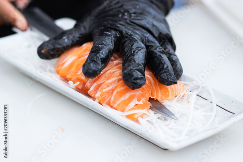 A chef wearing black glove using a knife to arrange fresh salmon sashimi on a white plate with shredded radish.