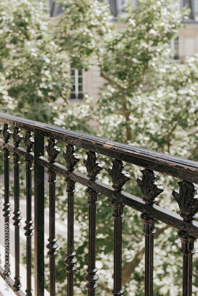 Fototapeta premium Classic Black Wrought Iron Balcony Railing with View of Green Trees, evocative european architecture, romantic old-world outdoor aesthetic, heritage french urban lifestyle.