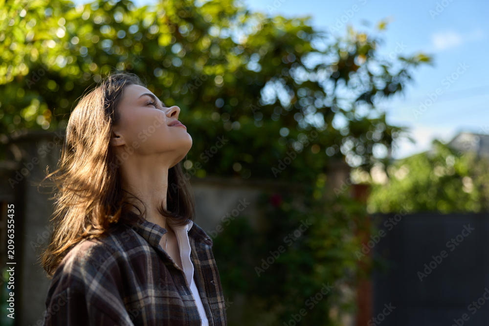 Fototapeta premium Smiling woman with shoulder length hair standing outdoors, head tilted toward the sun. Casual plaid shirt, nature backdrop, feeling relaxed, carefree, and reflective