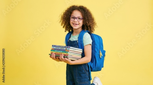Happy young girl with curly hair wearing glasses and a backpack holding a stack of school books on a yellow background