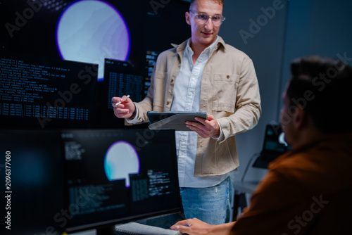 Bilde på lerret A focused blond cybersecurity analyst stands holding a tablet while briefing a colleague inside a dark security operations centre at night, defending the corporate network