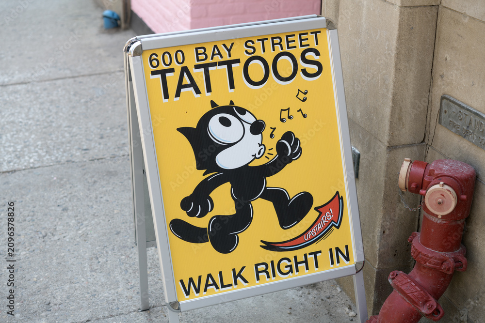 Fototapeta premium a-frame sidewalk sign outside 600 Bay St featuring a cartoon cat whistling towards a tattoo shop, Toronto
