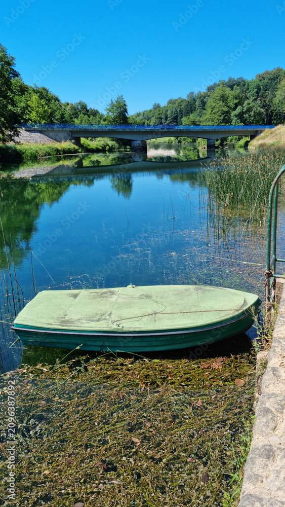 Fototapeta premium A small green rowboat is tied near the shore of the calm Dobra river in Jarce Polje with a concrete road bridge in the background under a blue sky